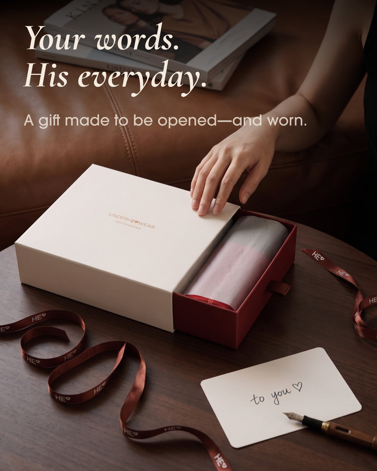 Hand opening an underherwear gift box beside a handwritten note and burgundy ribbon on a wooden table.
