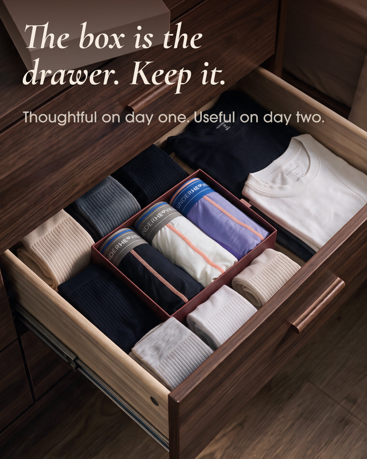 An underherwear box holding three rolled pairs of underwear in a wooden drawer beside folded socks and shirts.