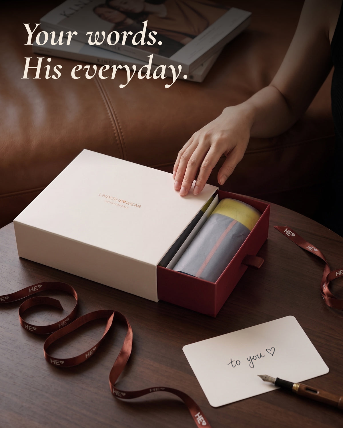 A hand opening an underherwear gift box beside a handwritten note and ribbon on a wooden table.